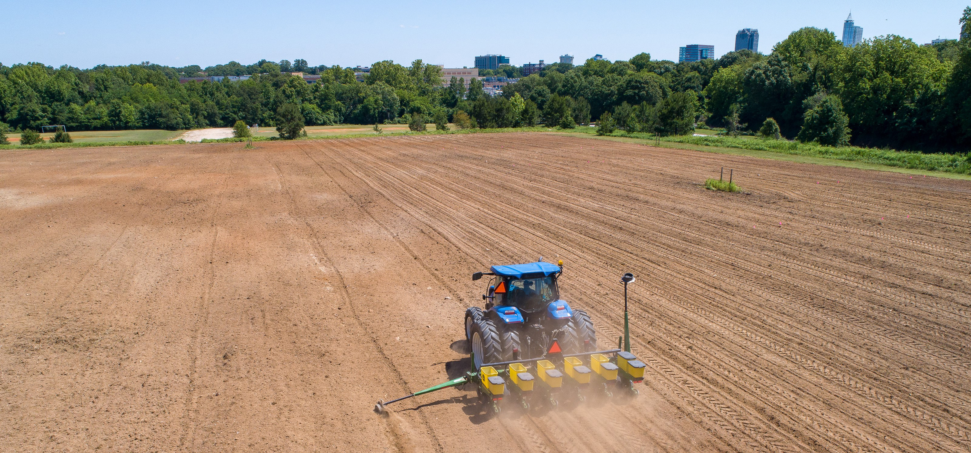 Aerial view of a tractor planting the sunflower field at Dix Park with the city skyline in the distance